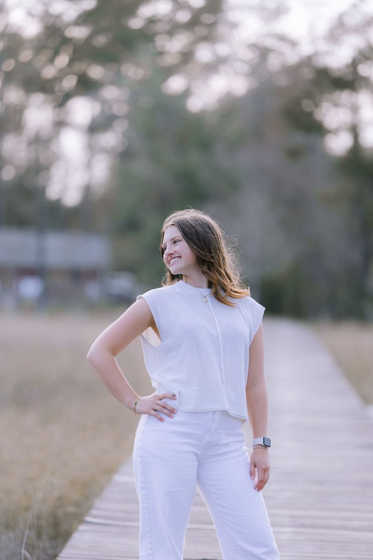 A senior in white standing on the boardwalk with a hand on her hip, looking off toward the pines