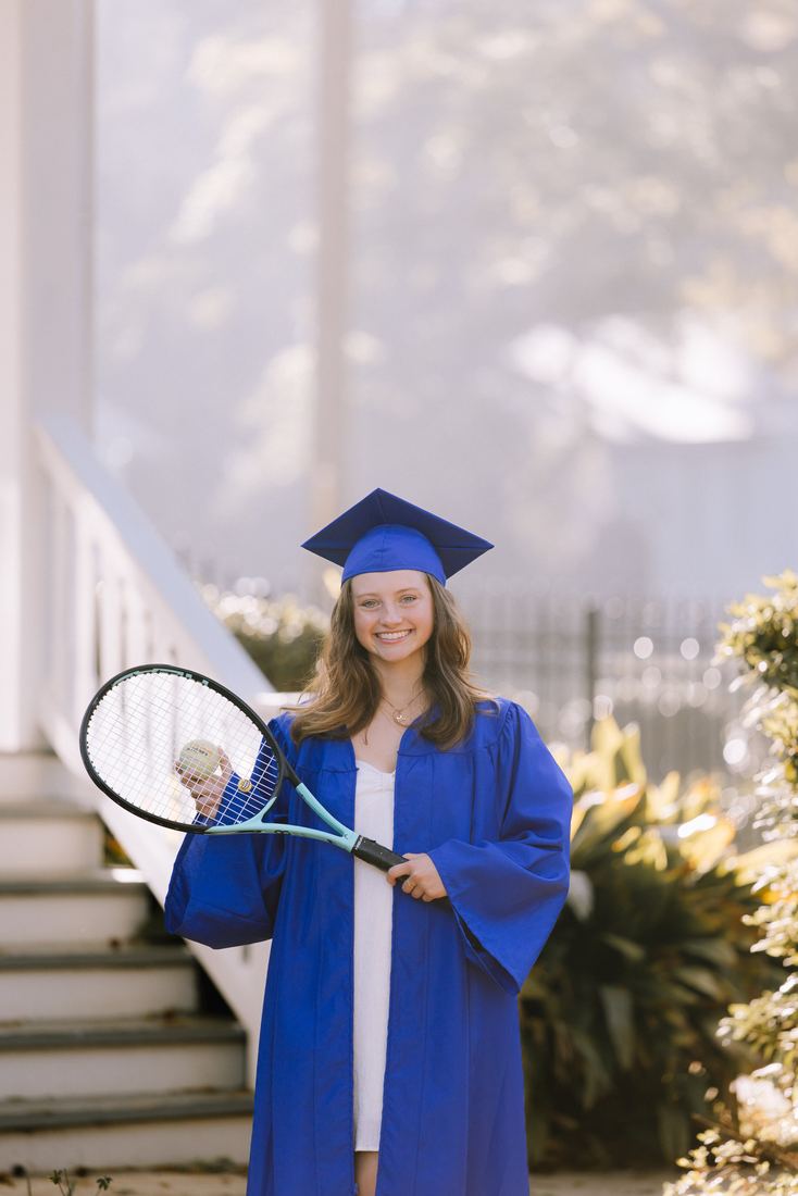 A graduate in a blue cap and gown holding a tennis racquet and ball beside a white staircase