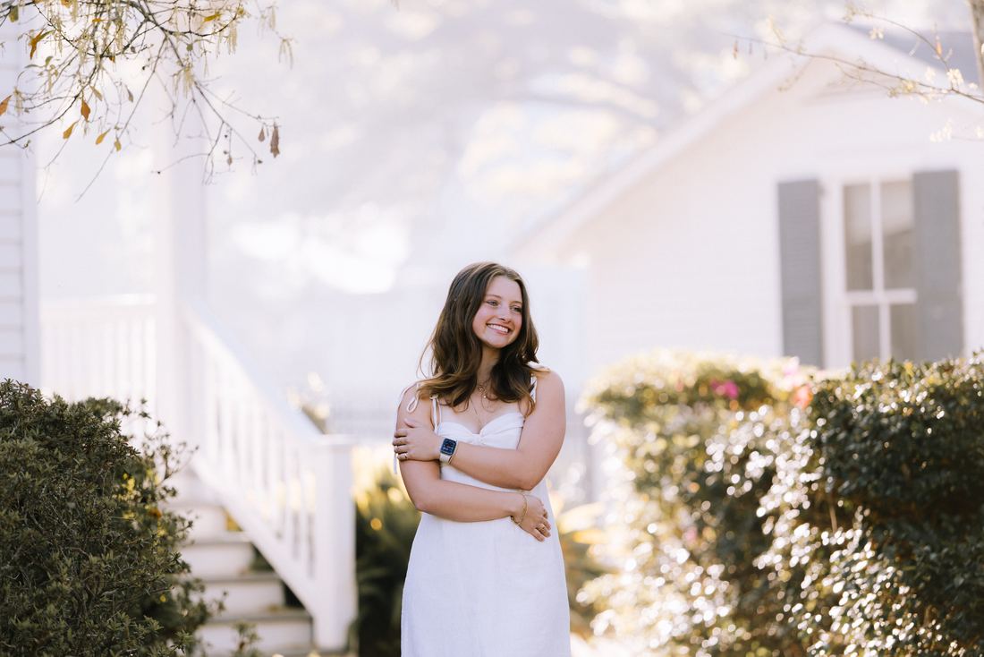 A senior in a white dress standing among azaleas in front of a white clapboard house, laughing off to the side