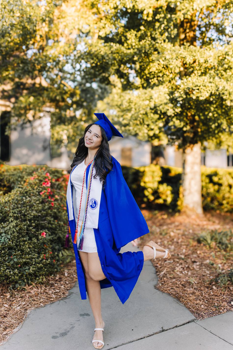 A senior in cap and gown with honor cords, kicking one foot up on a garden path