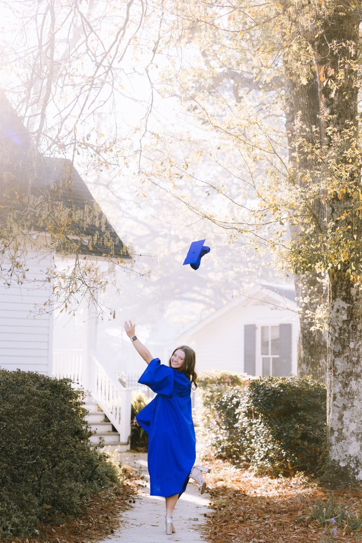 A graduate tossing her cap into the air on a sunlit sidewalk, looking back at the camera mid-throw