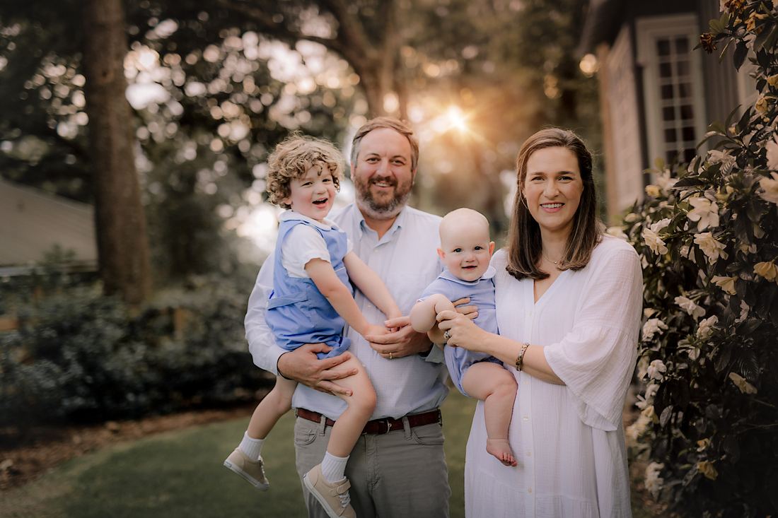 A family of four in a garden at golden hour, both boys in blue held by their parents, gardenias blooming beside them