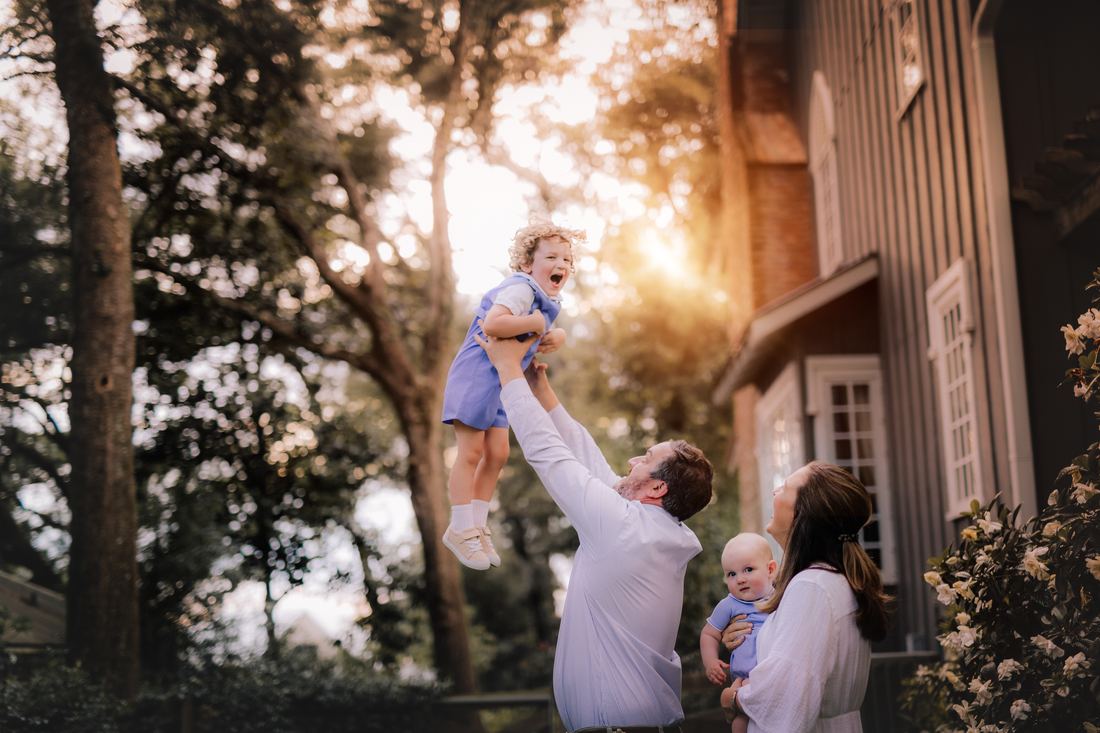 A father lifting his laughing son overhead beside the house while his wife looks up with the baby in her arms