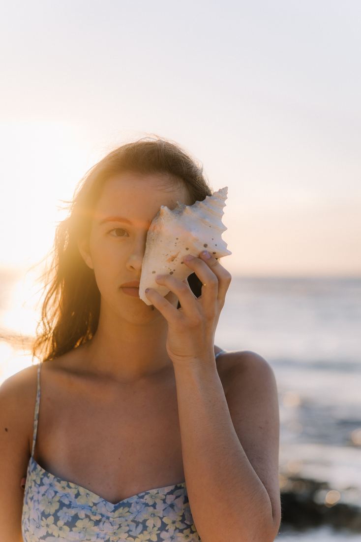 A senior at the shore holding a conch shell up over one eye, the low sun catching her hair