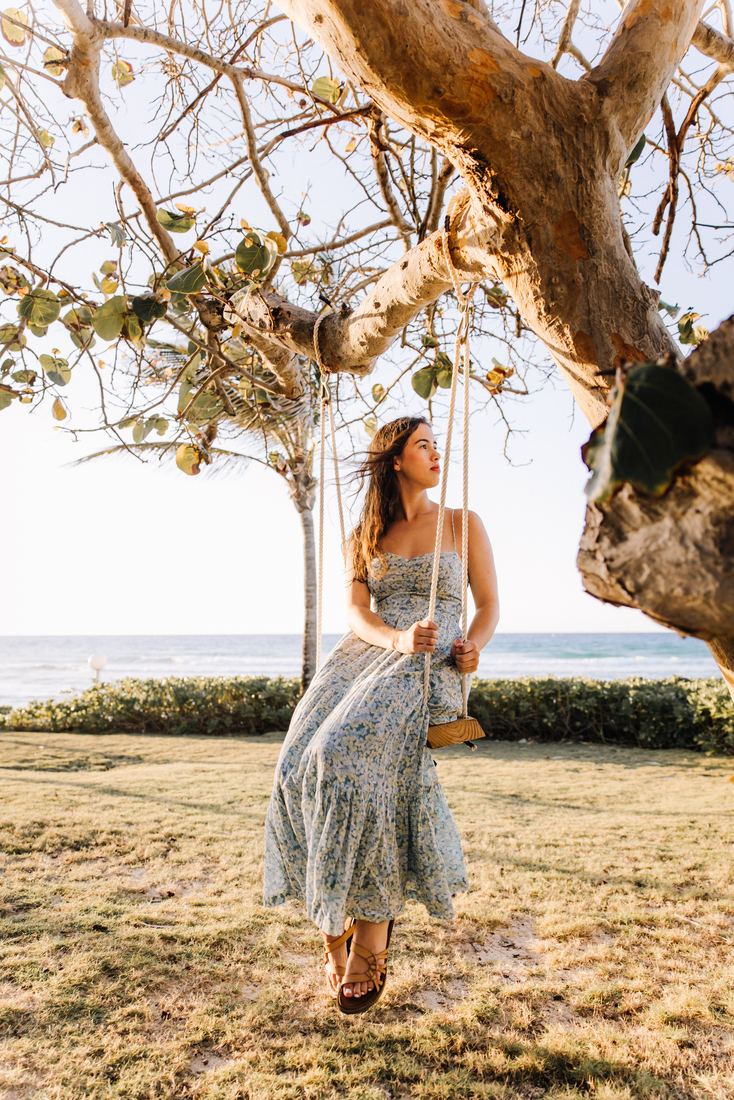 A senior in a blue floral dress sitting on a rope swing hung from a sea grape tree, the surf behind her
