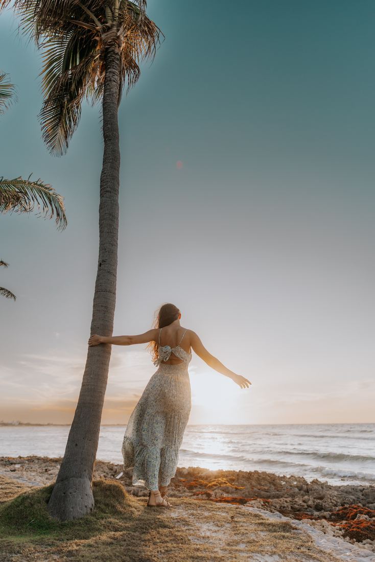 A senior leaning out from a palm trunk with one arm outstretched, facing the sunset over the water