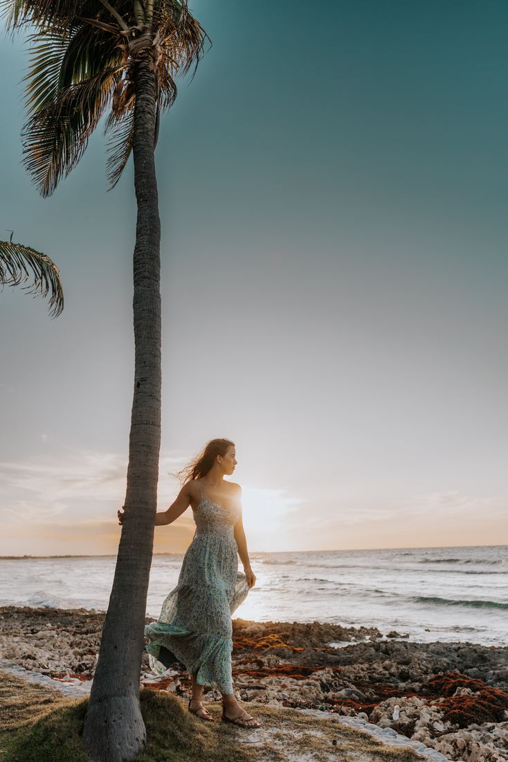 A senior standing at the base of a tall palm looking out to sea, her dress lifting in the wind