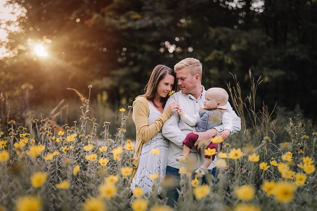 Parents standing close in a field of yellow wildflowers, their baby on his father's hip reaching for the blossom in his mother's hand
