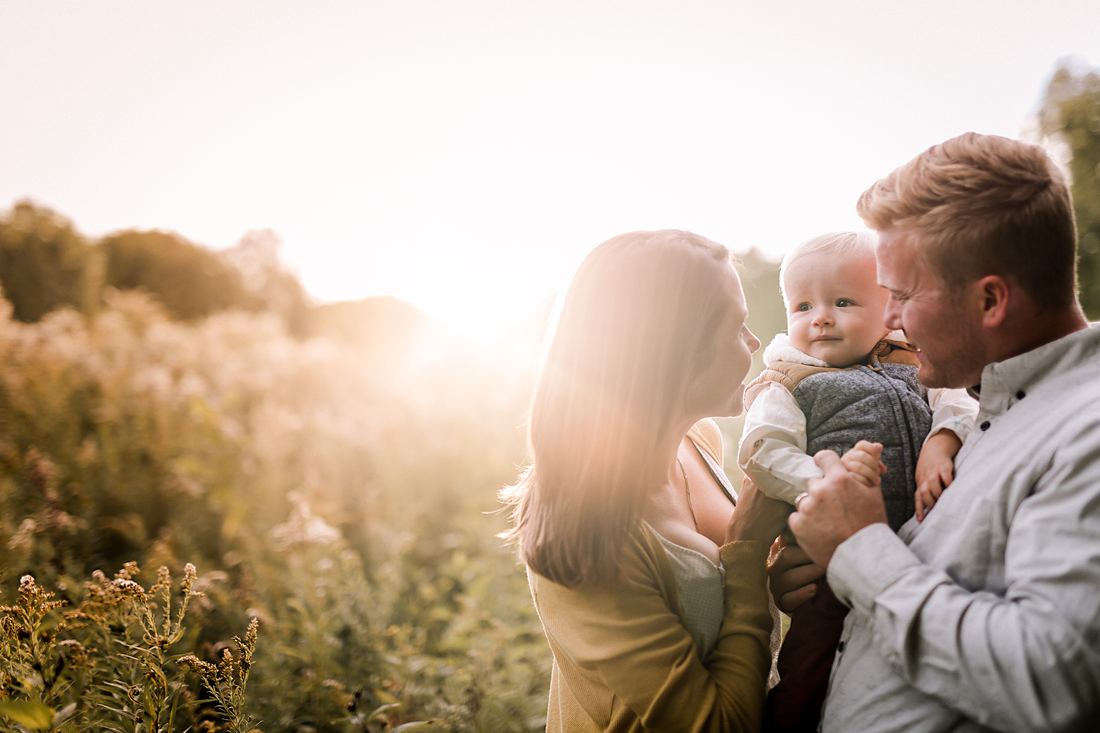 A backlit close view of parents leaning toward each other with their baby held between them, the field washed out in sun