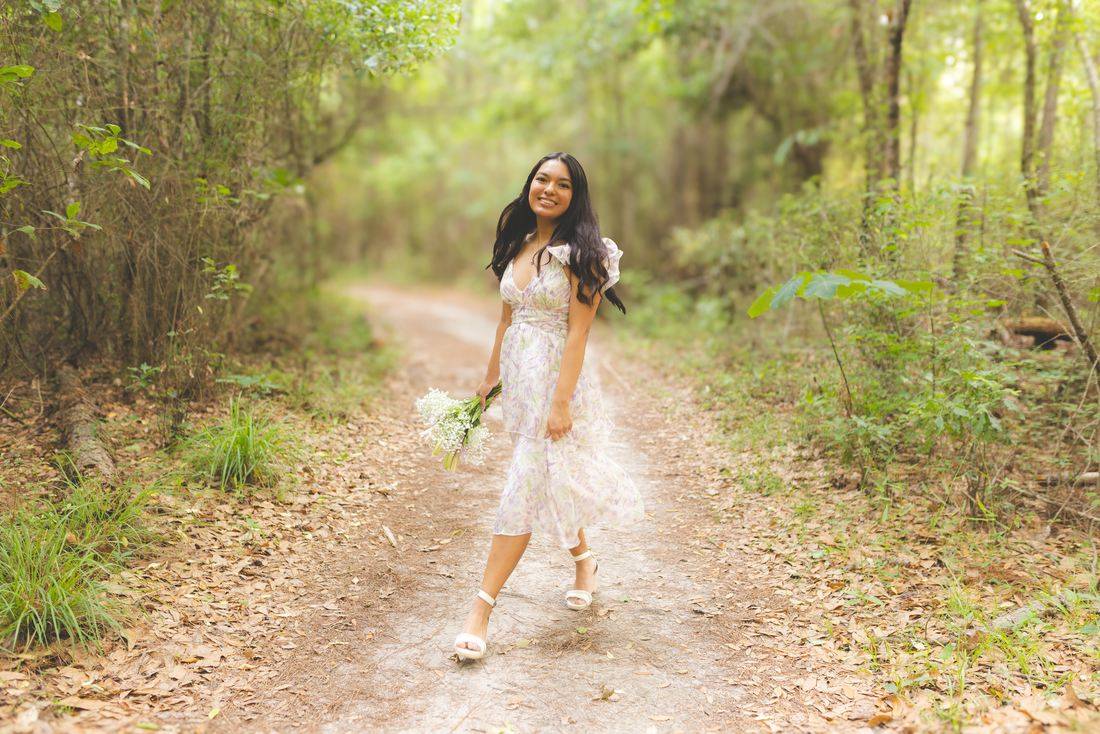 A senior walking a leafy trail in a white dress, holding a bouquet