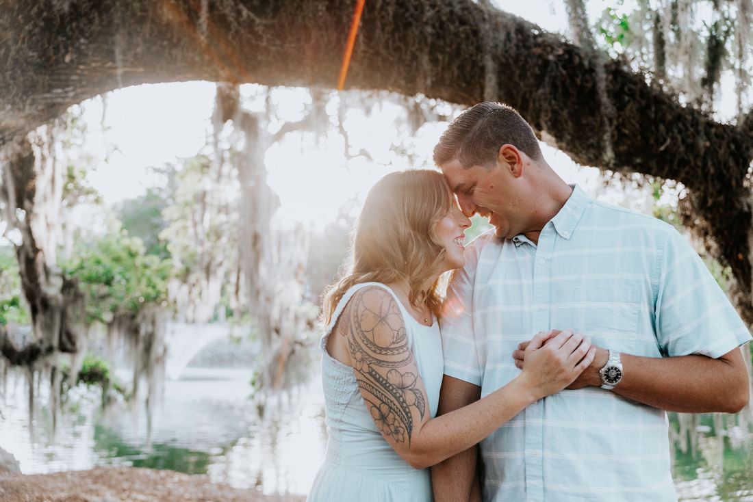 A couple laughing forehead to forehead beneath a mossy live oak beside the water