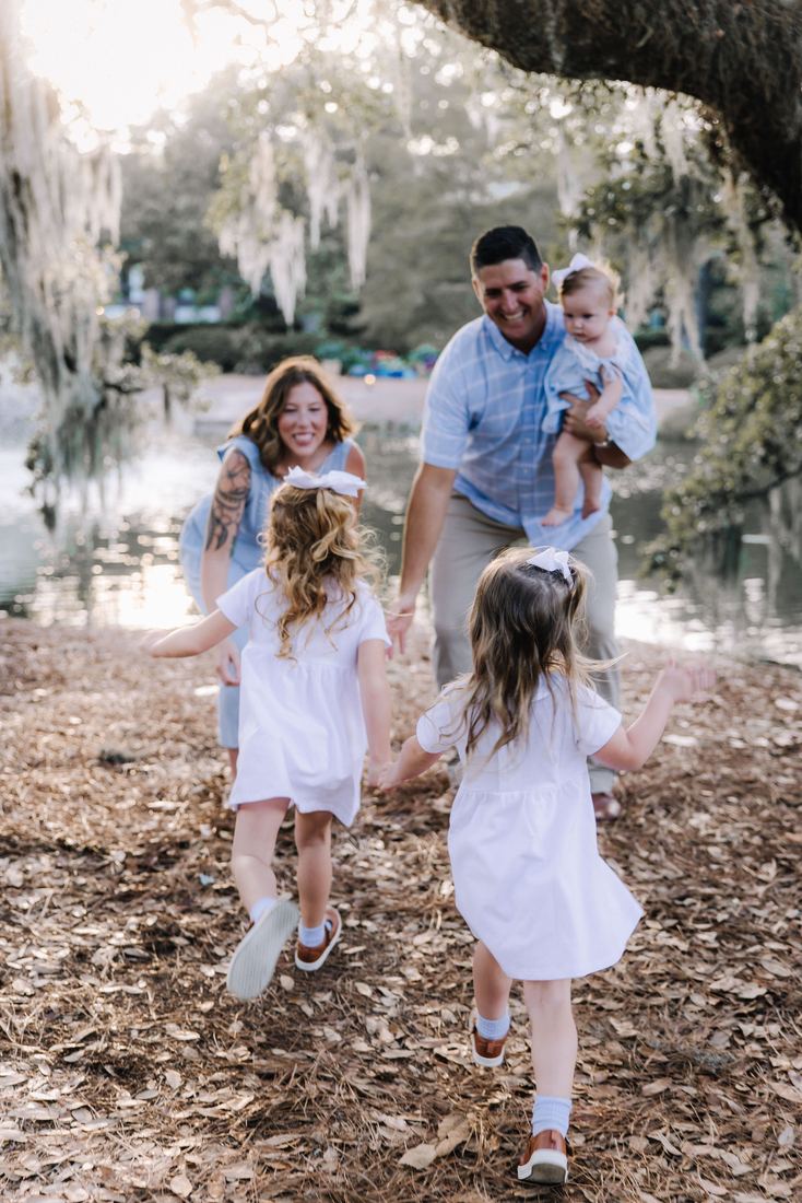 Two girls in white dresses running hand in hand toward their parents under the live oaks, the baby on her father's hip
