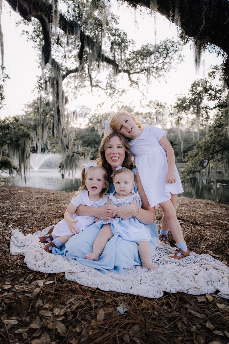 A mother seated on a lace blanket beneath a moss-draped oak with her four girls gathered around her, the fountain on the water behind them