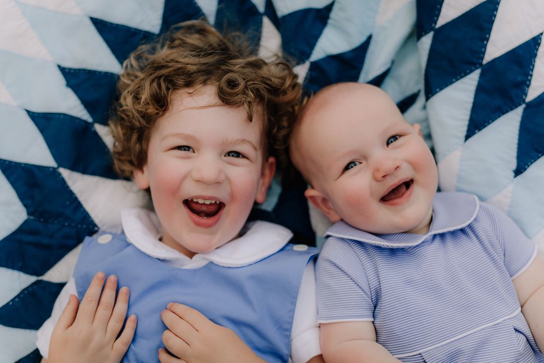 Overhead view of two brothers lying side by side on a navy and white quilt, both laughing up at the camera