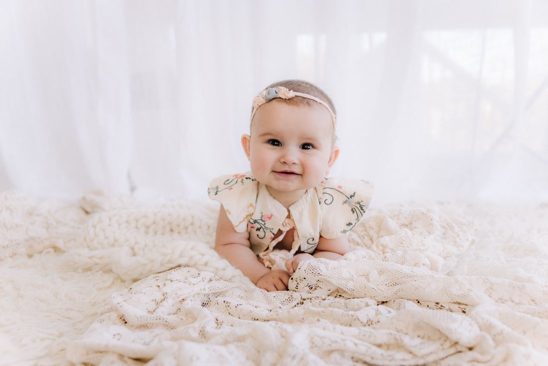A baby in an embroidered collar and blush headband pushing up on her arms on a cream lace blanket