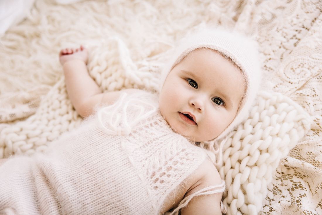 A baby in a knit bonnet and pale pink romper lying on a chunky cream blanket, looking up at the camera