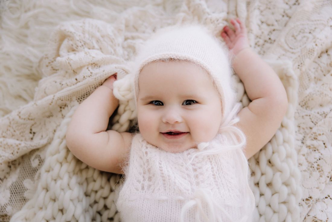 Overhead view of a baby in a soft knit bonnet with both arms flung above her head, grinning