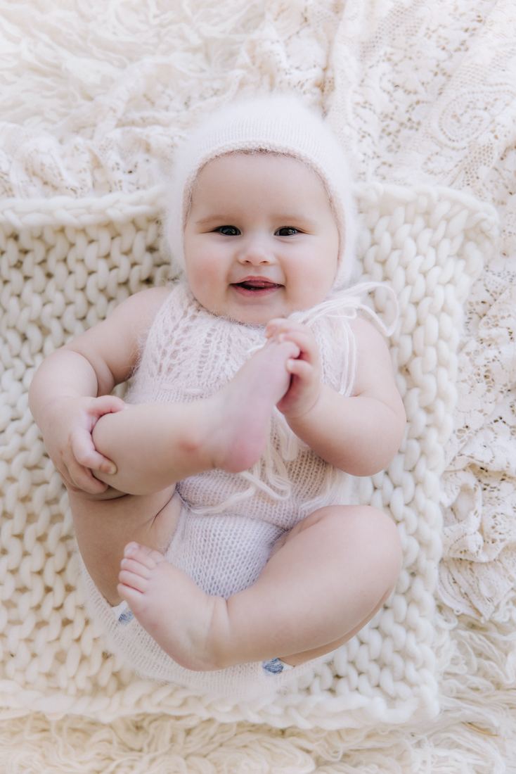 A baby in a knit bonnet on a cream blanket, holding onto one foot with both hands