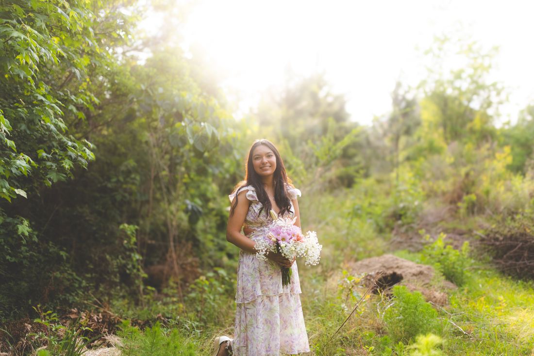 A senior in a floral dress with a bouquet, backlit by sun through the trees