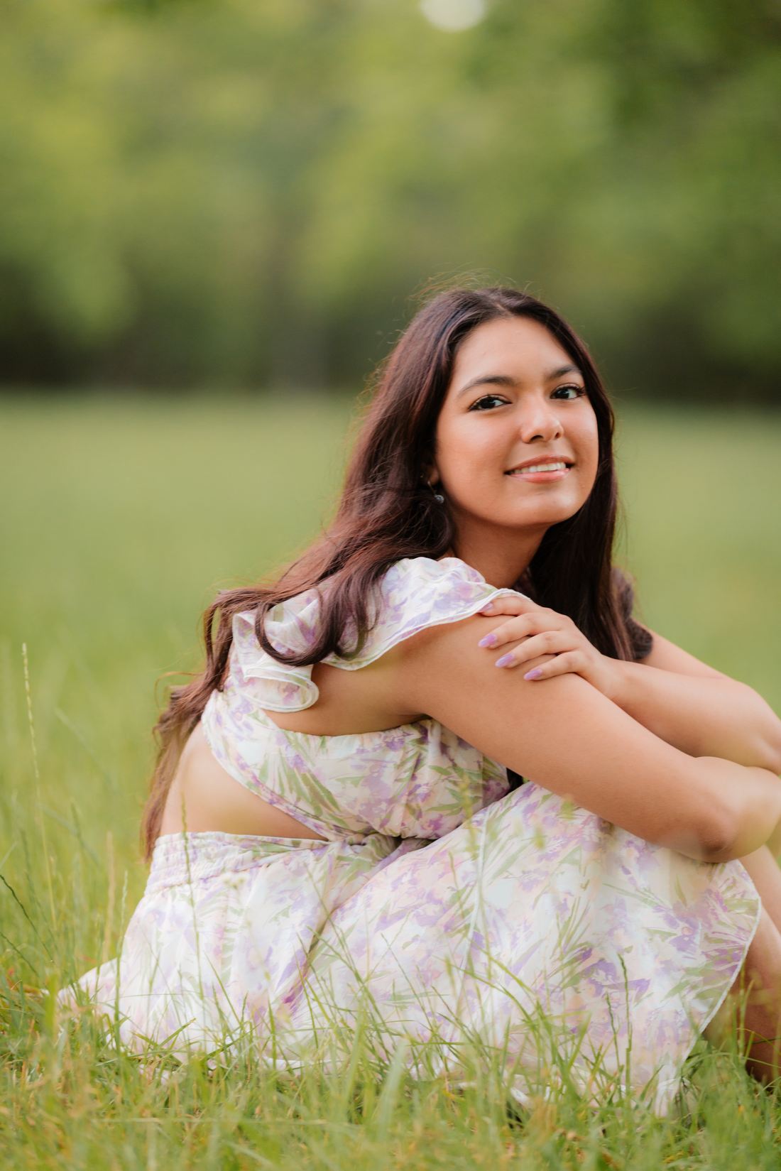 A senior sitting in tall grass with her arms around her knees, smiling
