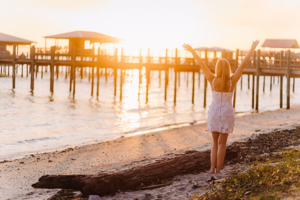 A senior on the shore with her arms raised toward the sun setting behind the pier