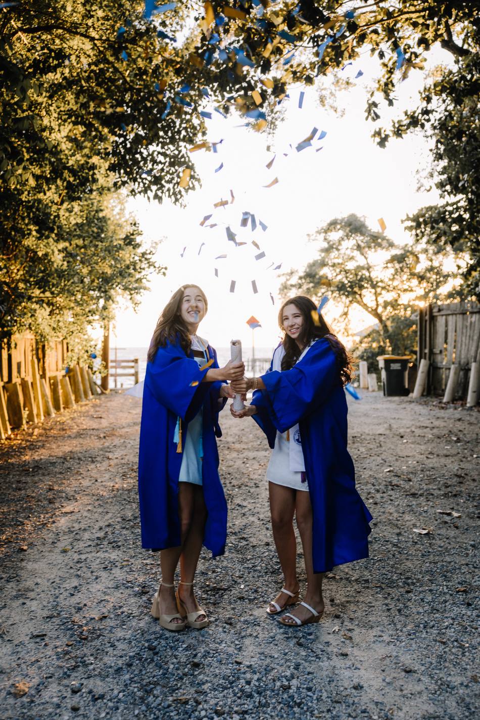 Two graduates in blue gowns popping confetti under the oaks