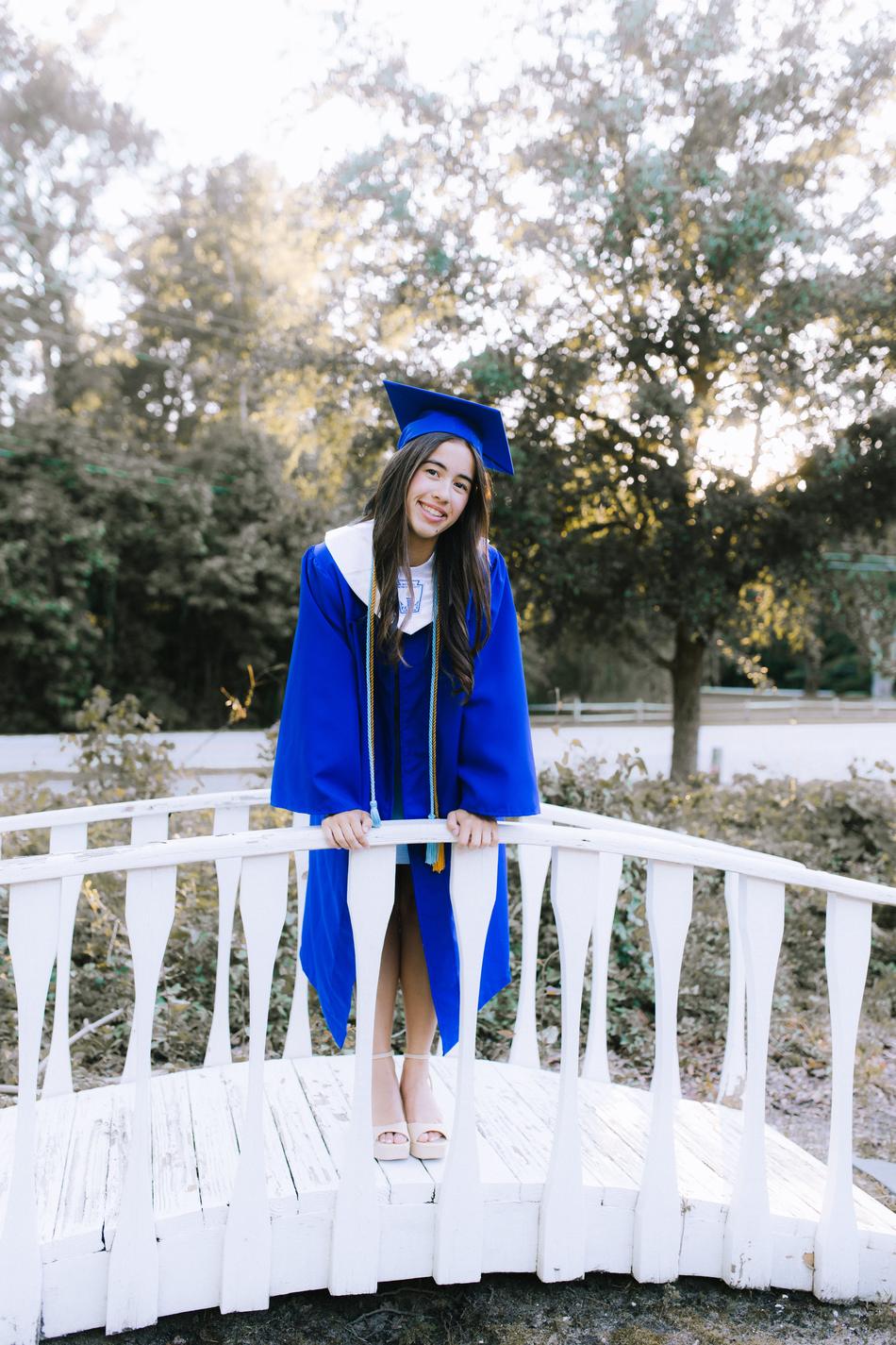A graduate in cap and gown leaning on the rail of a white garden bridge