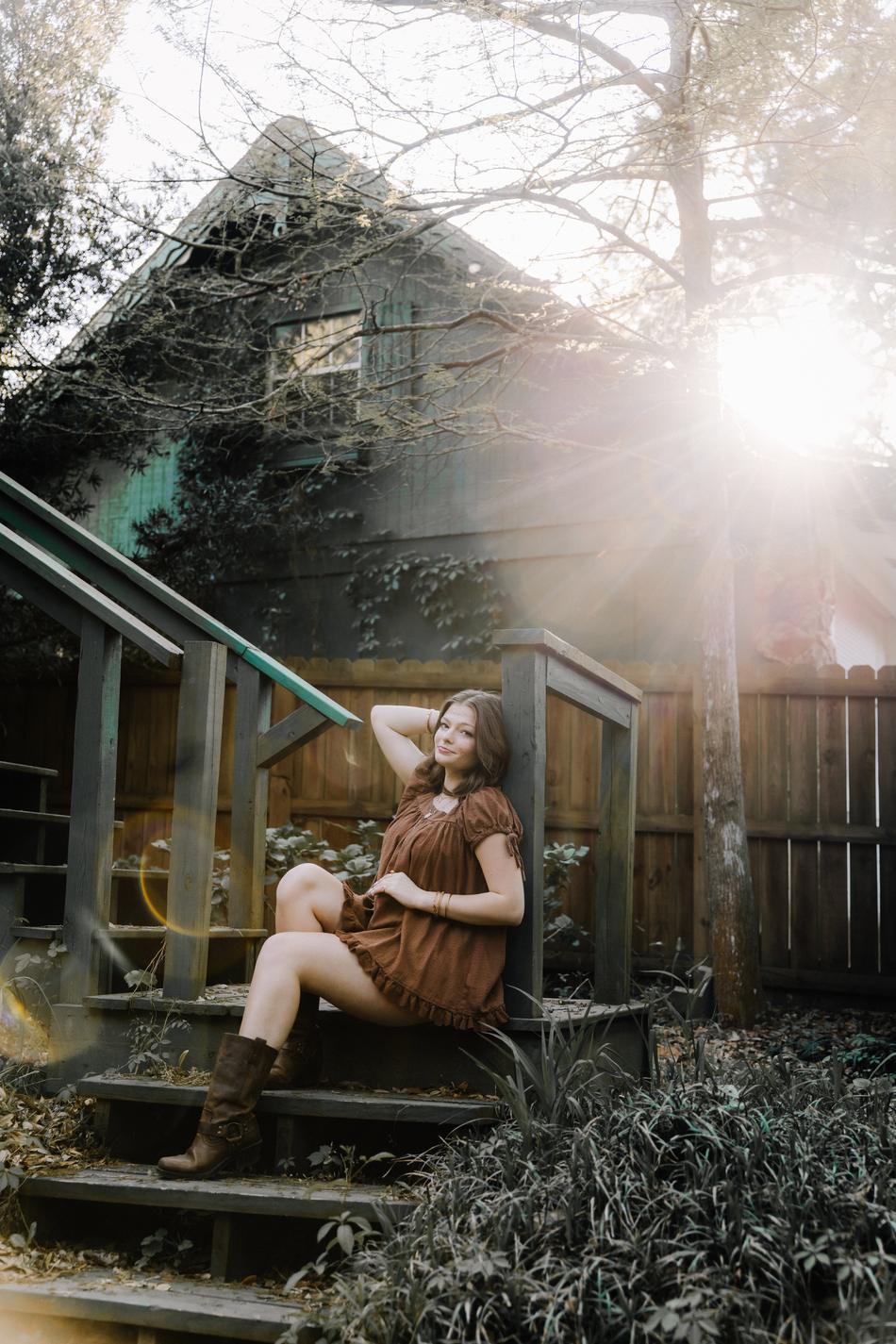 A senior in a rust dress and boots on weathered porch steps with the sun flaring behind