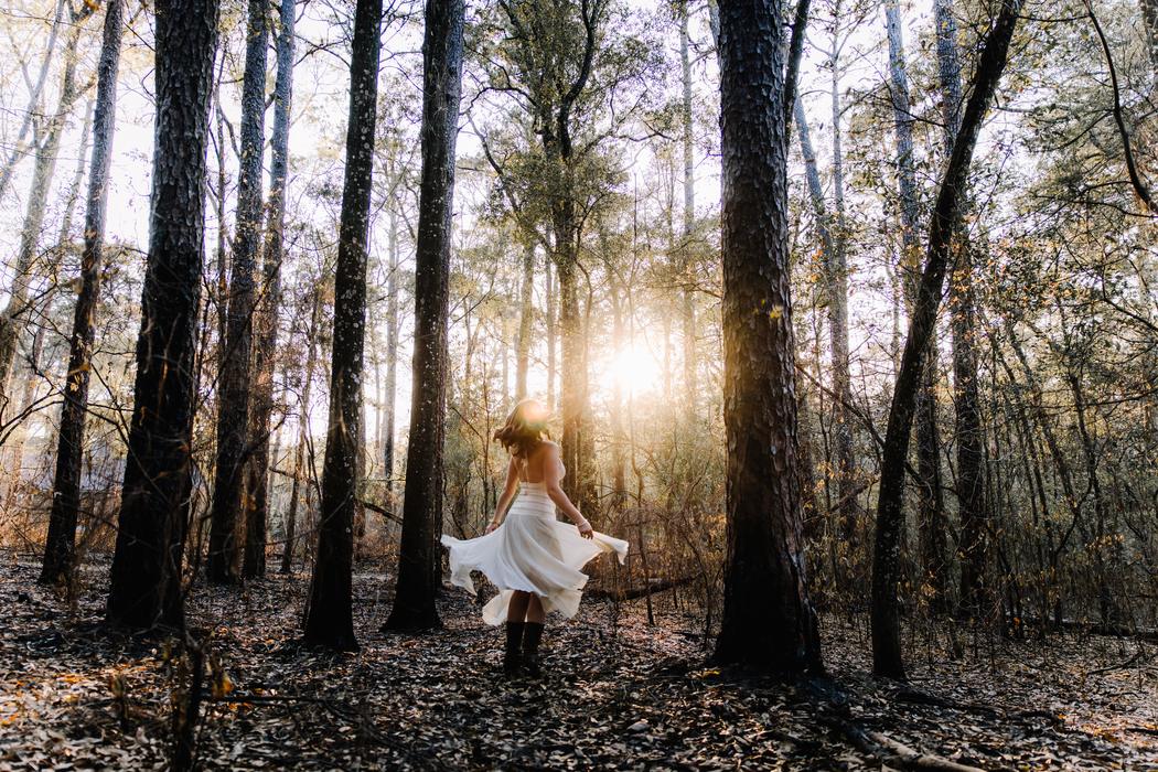 A senior dancing in a clearing among tall pines with the sun low behind the trunks