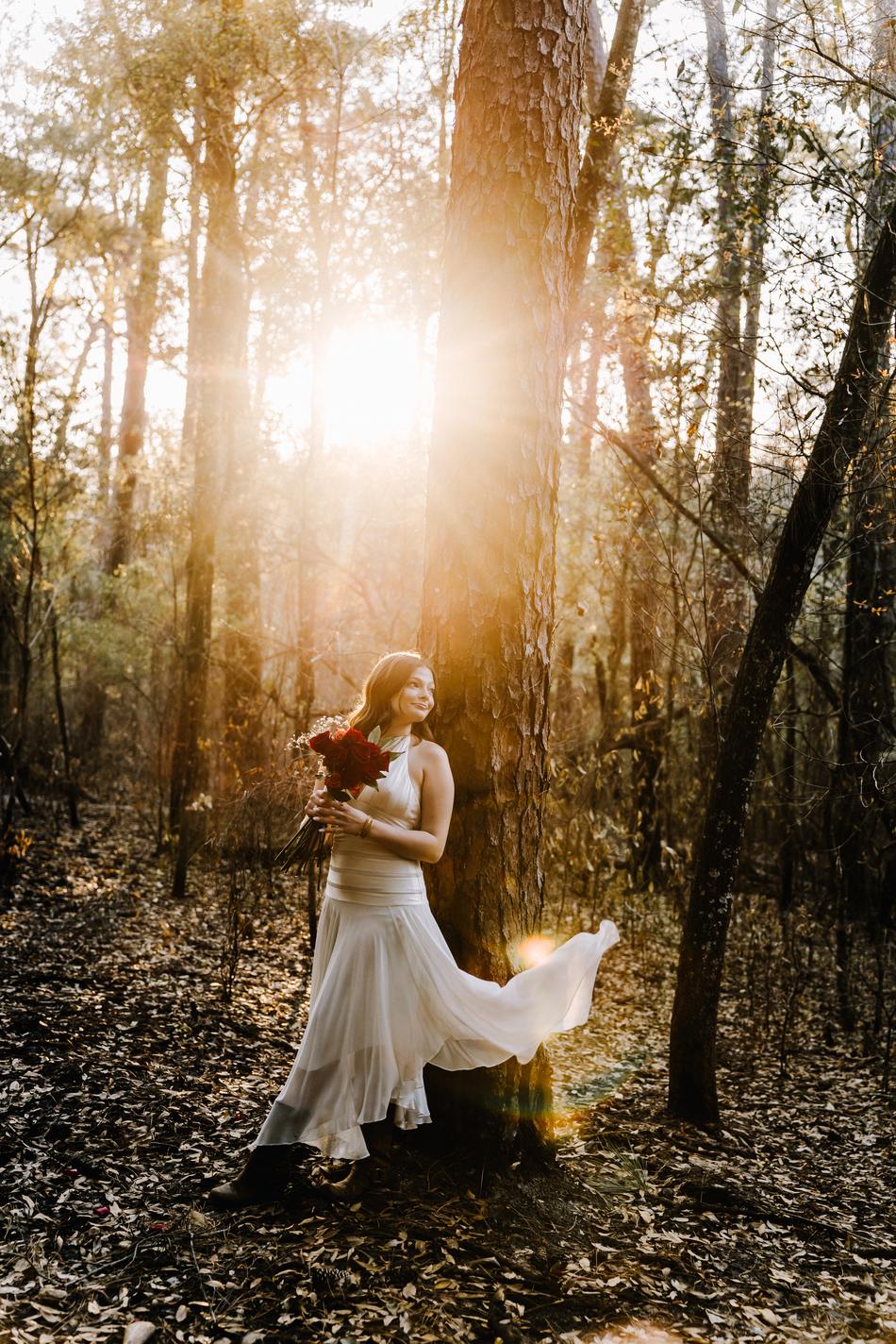 A senior in a white gown standing in the woods, backlit by the setting sun