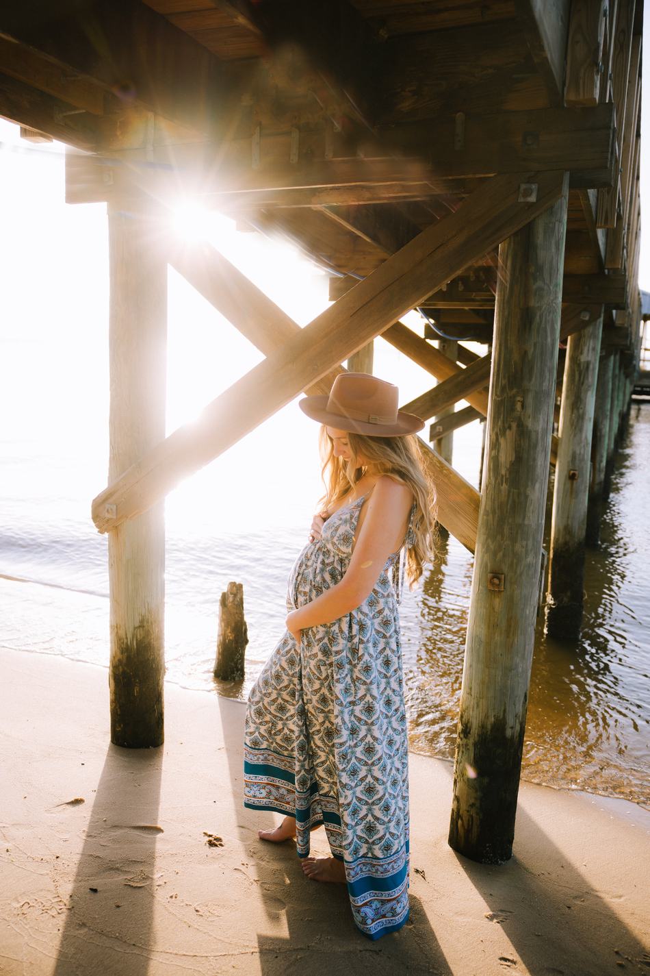 An expecting mother in a patterned dress and wide-brimmed hat beneath the pier