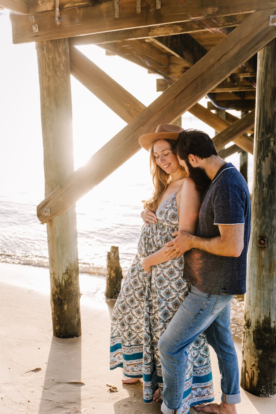 An expecting couple holding her belly together in the sand under the pier