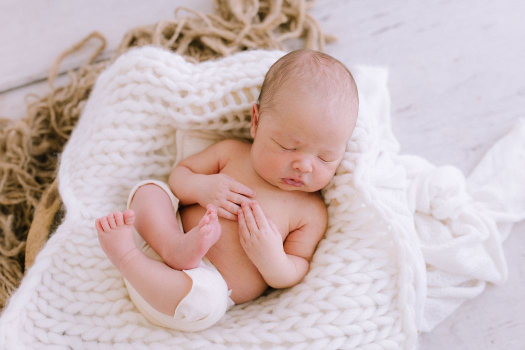 A newborn curled up asleep on a chunky cream knit blanket