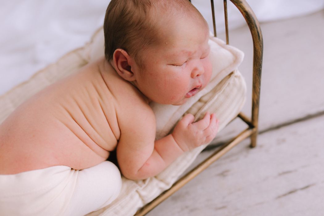 A newborn asleep on a tiny brass bed with a linen pillow