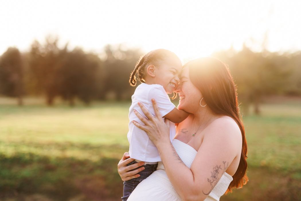 An expecting mother lifting her little one, the two of them laughing forehead to forehead at sunset