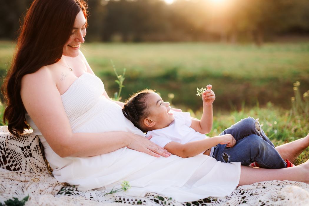 A mother and her child resting against her belly in the grass, holding a wildflower