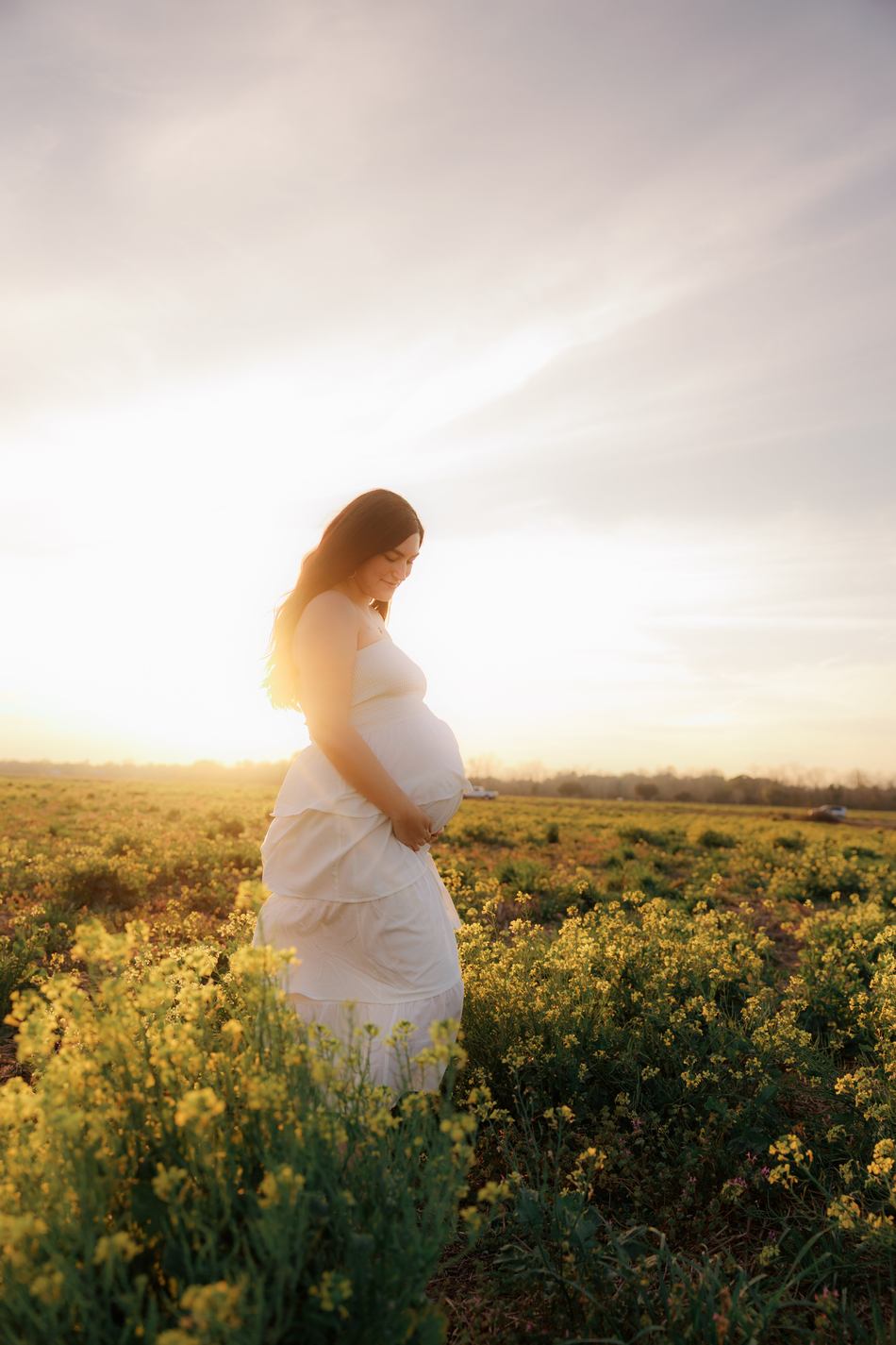 An expecting mother in a white dress standing in a field of yellow blooms at golden hour