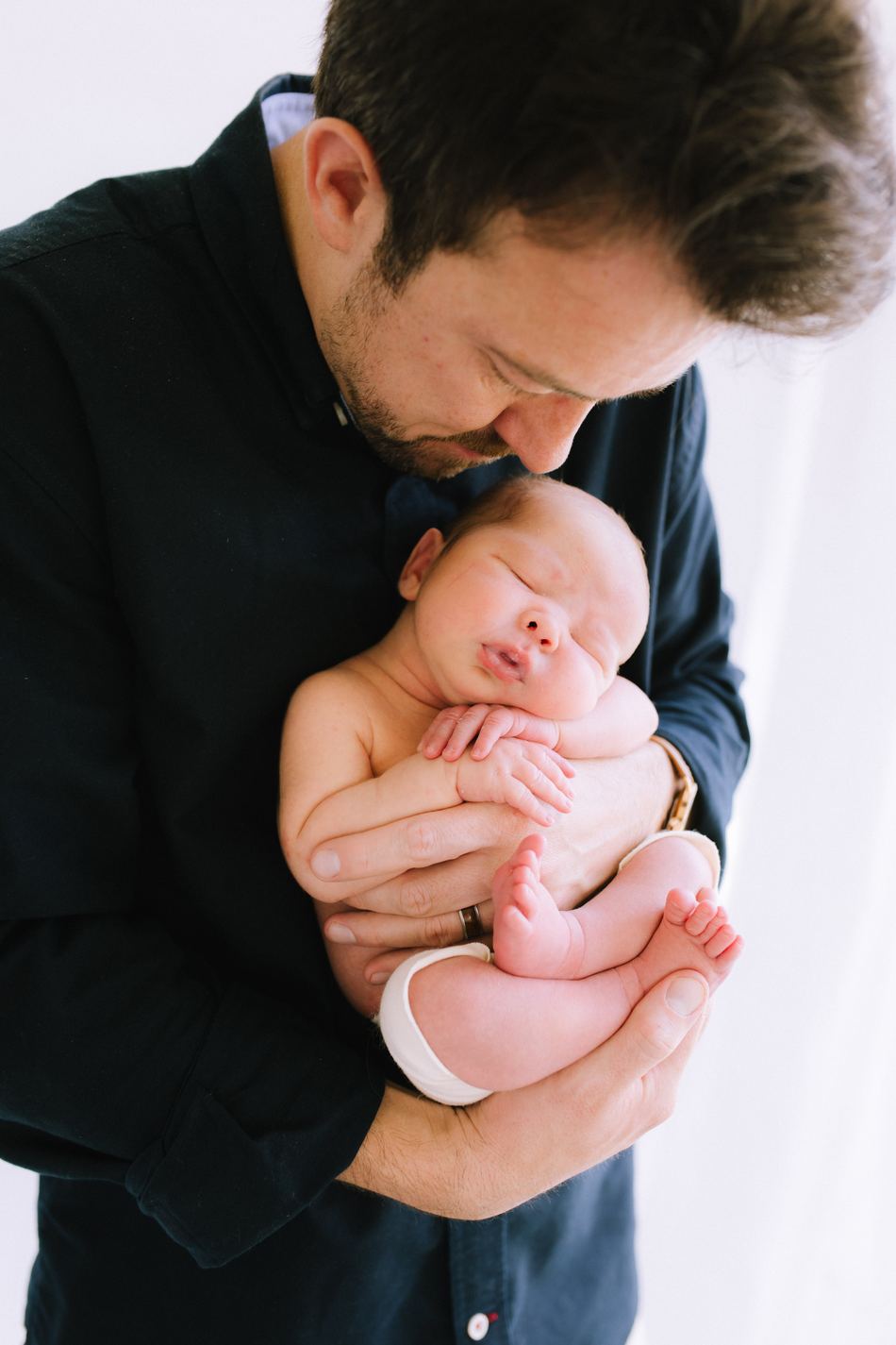 A father cradling his sleeping newborn against his chest in soft window light