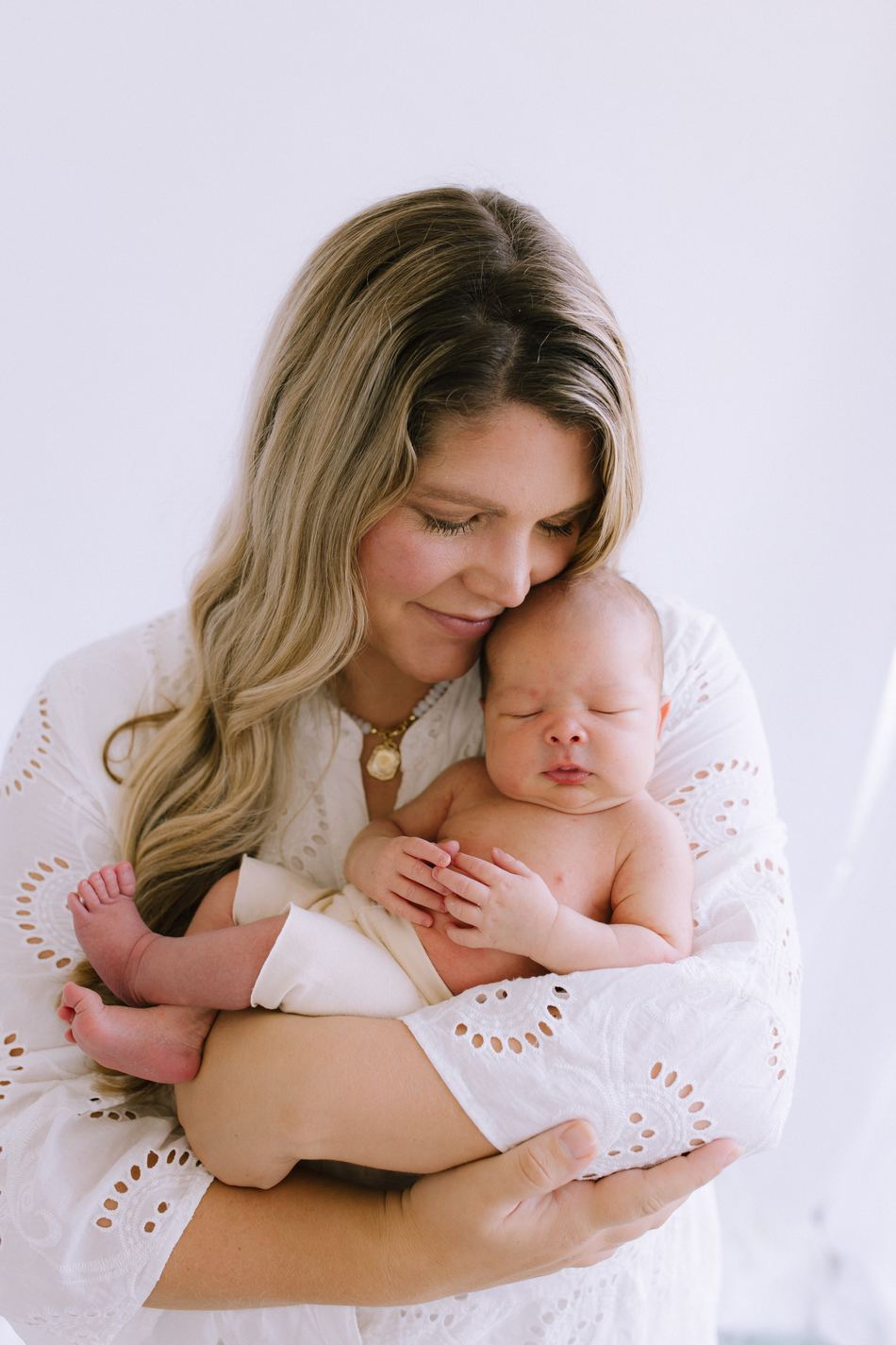 A mother in white eyelet resting her cheek against her sleeping newborn