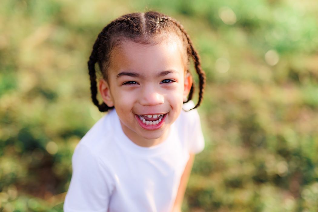 A little girl in braids and a white tee grinning wide in a sunlit field