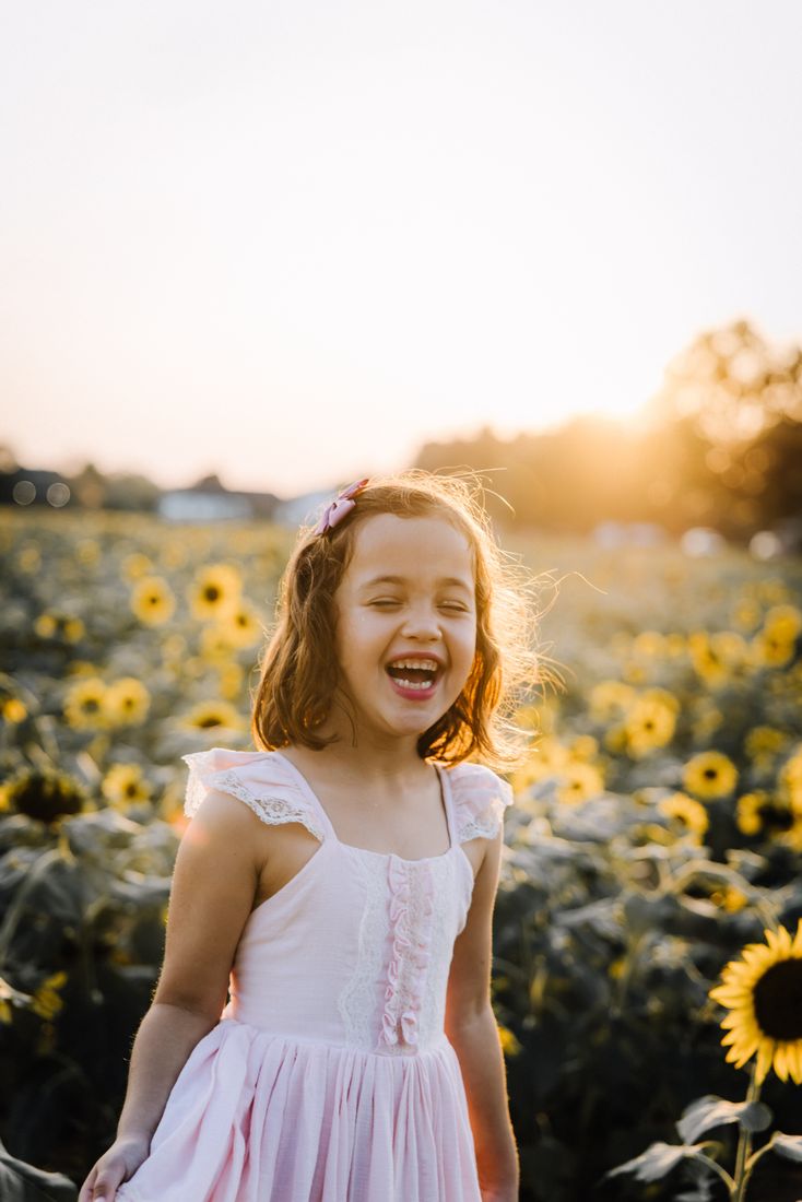 A girl in a pink lace dress laughing with her eyes closed in a field of sunflowers