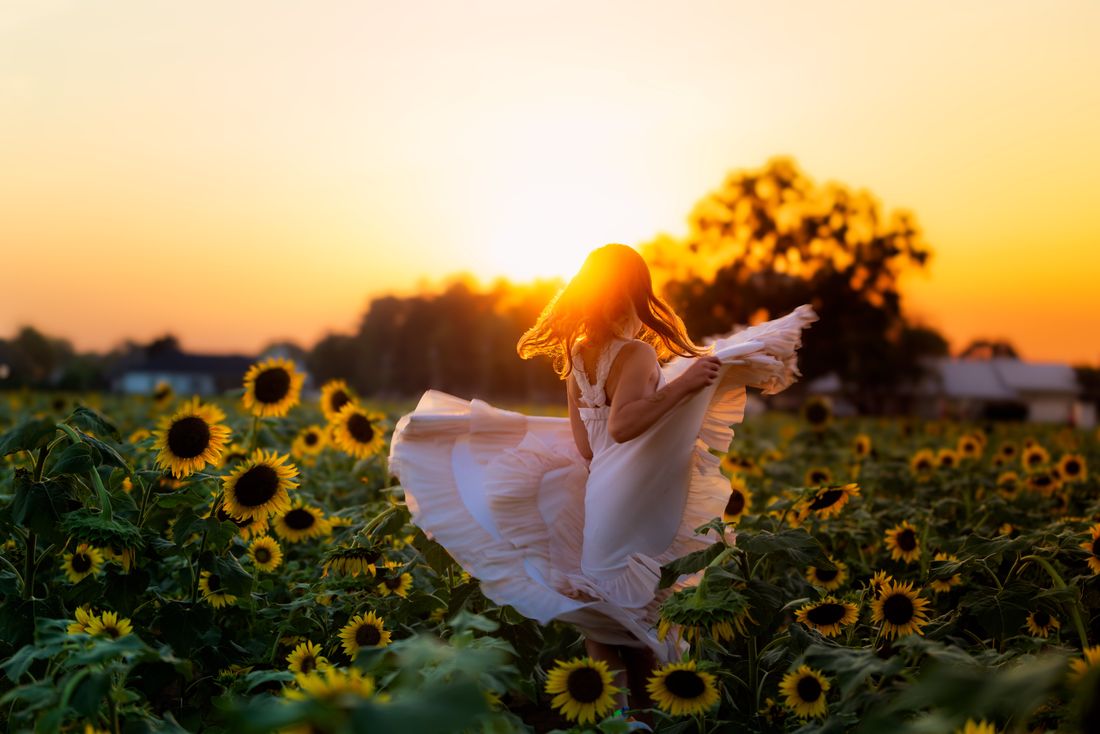 A girl twirling her white dress in a sunflower field as the sun drops behind the trees