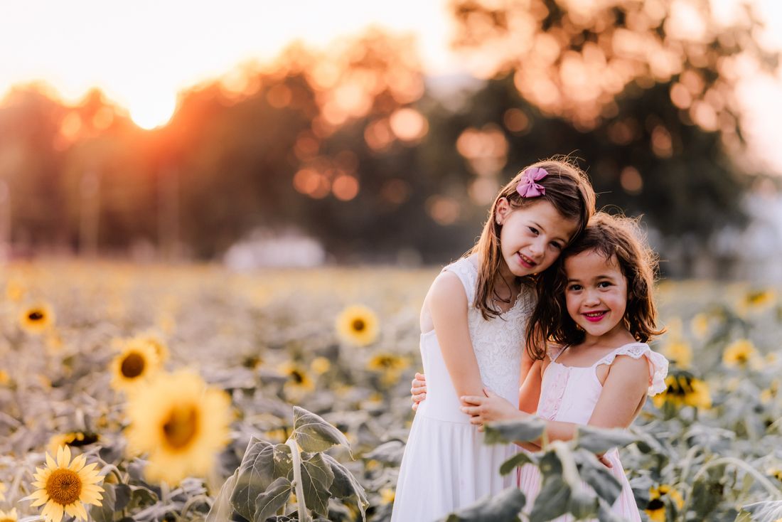 Two sisters hugging and smiling together in a field of sunflowers at golden hour