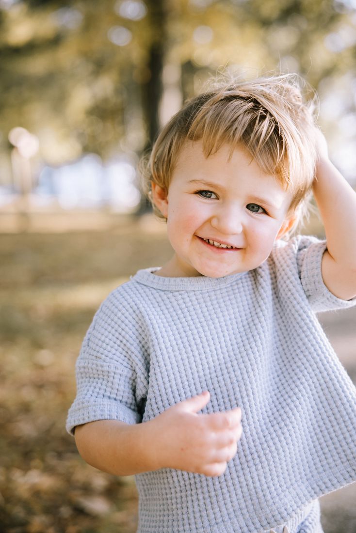 A toddler in a grey waffle tee grinning with a hand in his hair among the autumn trees