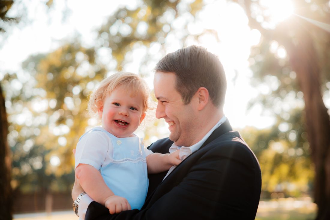 A father in a navy blazer laughing with his toddler son in his arms beneath the trees