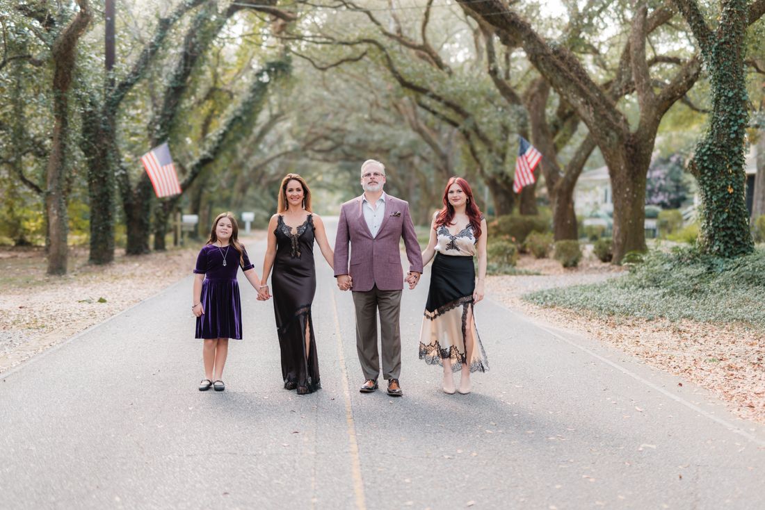 A family of four holding hands down the middle of an oak-lined street, dressed up for the evening