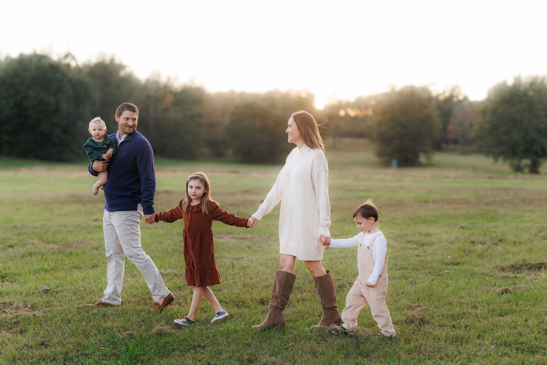 A family of five walking hand in hand across an open field at sunset, the baby on her father's hip