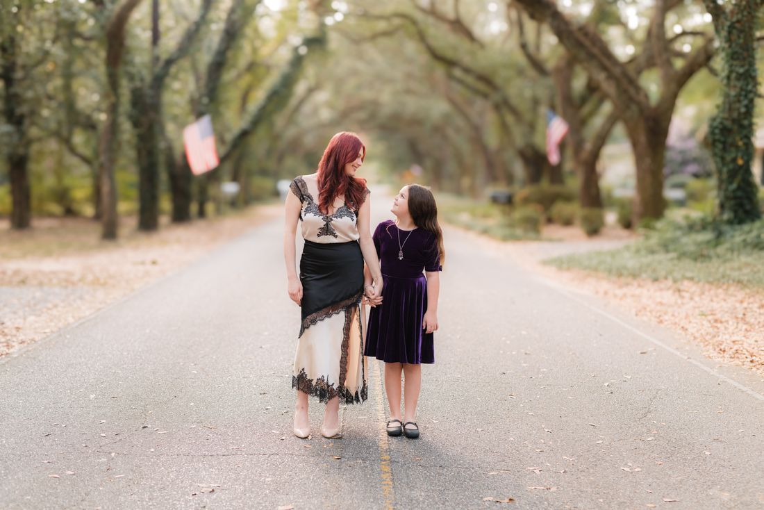 An older sister and her little sister walking hand in hand down an oak-lined street, looking at each other