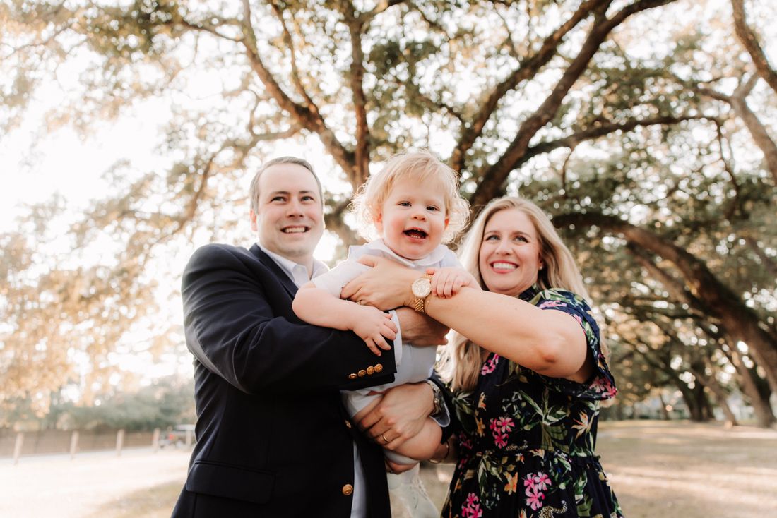 Parents holding their laughing toddler up between them under the live oaks