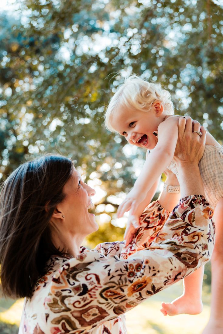 A mother lifting her toddler overhead in the dappled light beneath an oak